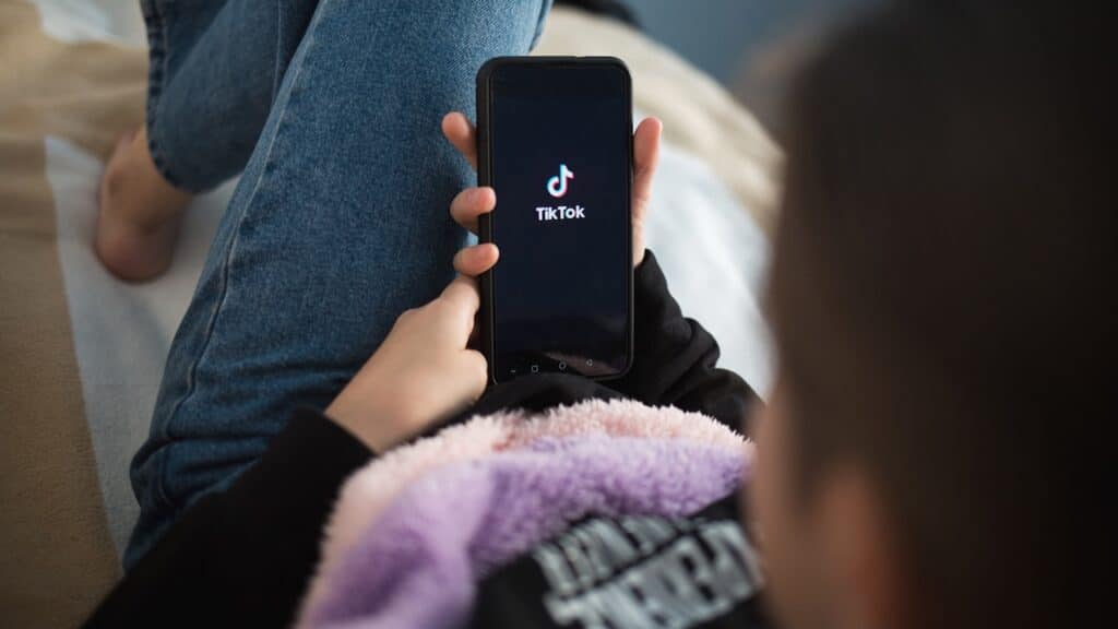 child with phone in his hands is lying at home on bed. Social Media tiktok is loaded on screen.