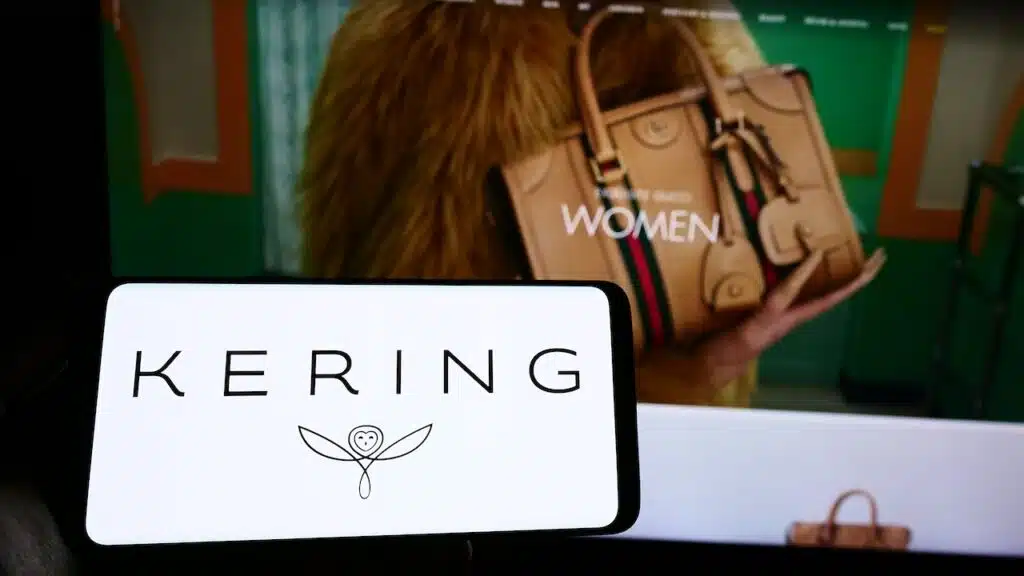 Person holding smartphone with logo of French luxury goods company Kering S.A. on screen in front of website.