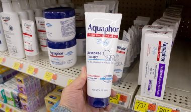 A view of a hand holding a container of Aquaphor baby healing ointment, on display at a local retail store.