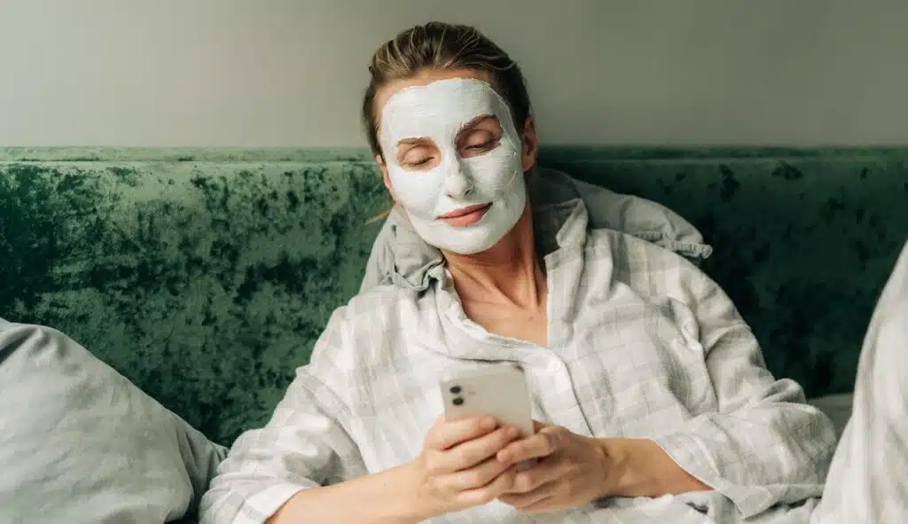 Young woman lying in bed with a clay mask on her face uses a mobile phone to surf the internet.