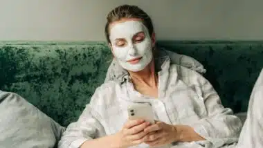 Young woman lying in bed with a clay mask on her face uses a mobile phone to surf the internet.