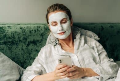 Young woman lying in bed with a clay mask on her face uses a mobile phone to surf the internet.