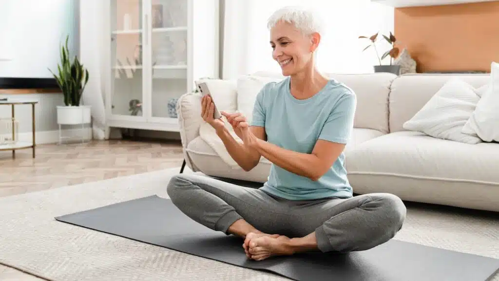 Middle-aged flexible woman in fitness outfit sitting in lotus position while using cellphone for sport app.