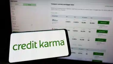 Person holding mobile phone with logo of American financial company Credit Karma Inc. on screen in front of web page.