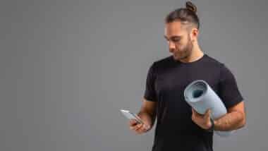 A man standing and holding a yoga mat in one hand and a cell phone in the other.