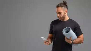 A man standing and holding a yoga mat in one hand and a cell phone in the other.
