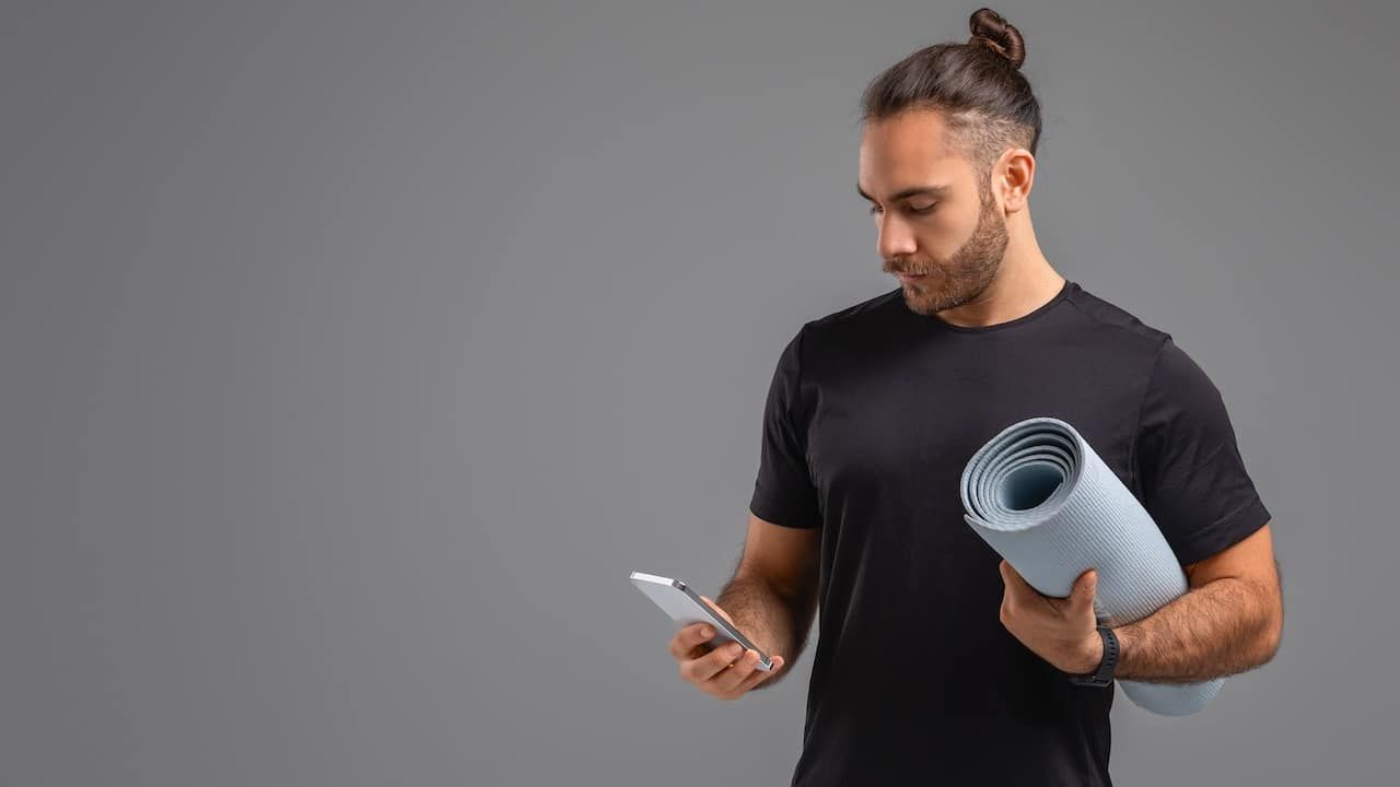 A man standing and holding a yoga mat in one hand and a cell phone in the other.