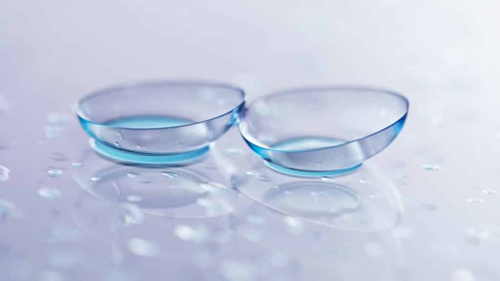 Pair of contact lenses and water drops on light mirror surface, closeup