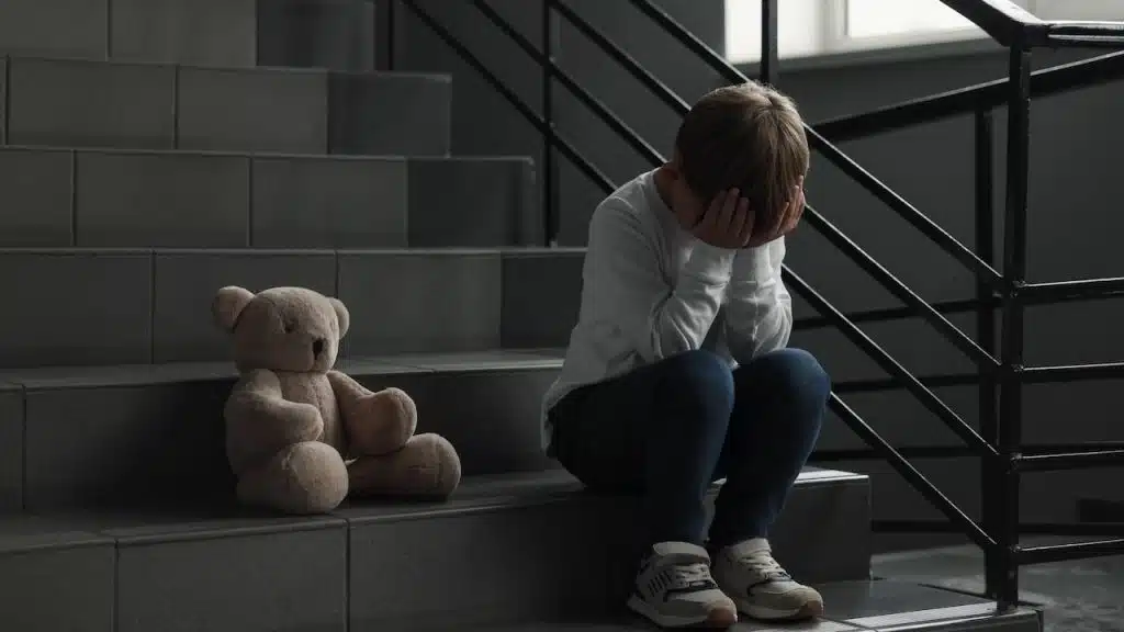 Upset little boy with toy bear on stairs indoors