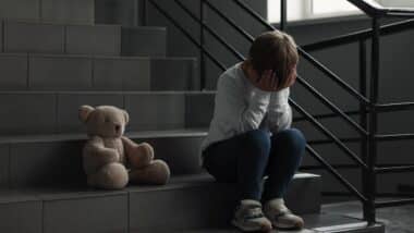 Upset little boy with toy bear on stairs indoors