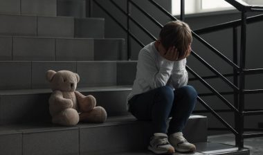 Upset little boy with toy bear on stairs indoors
