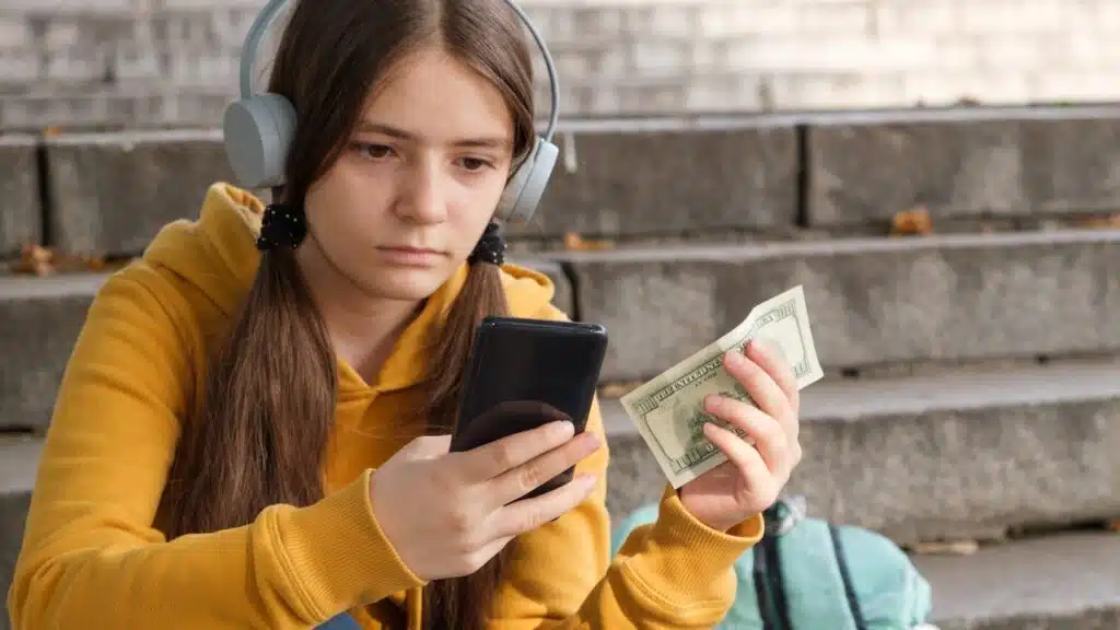 The teenager holds dollar bills and looks at the smartphone screen.