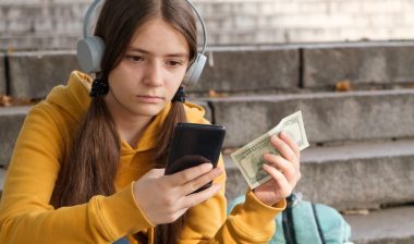 The teenager holds dollar bills and looks at the smartphone screen.