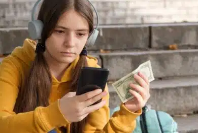 The teenager holds dollar bills and looks at the smartphone screen.
