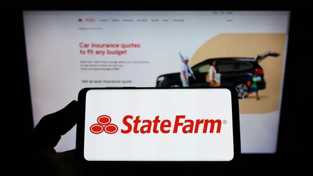Person holding smartphone with logo of State Farm Mutual Automobile Insurance Company on screen in front of website.