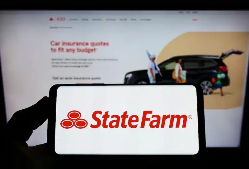 Person holding smartphone with logo of State Farm Mutual Automobile Insurance Company on screen in front of website.