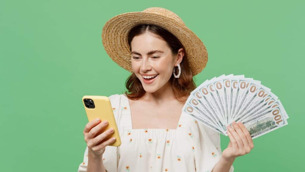 Young happy smiling woman she 20s in white dress hat hold in hand use mobile cell phone fan of cash money in dollar banknotes isolated on plain pastel light green background.
