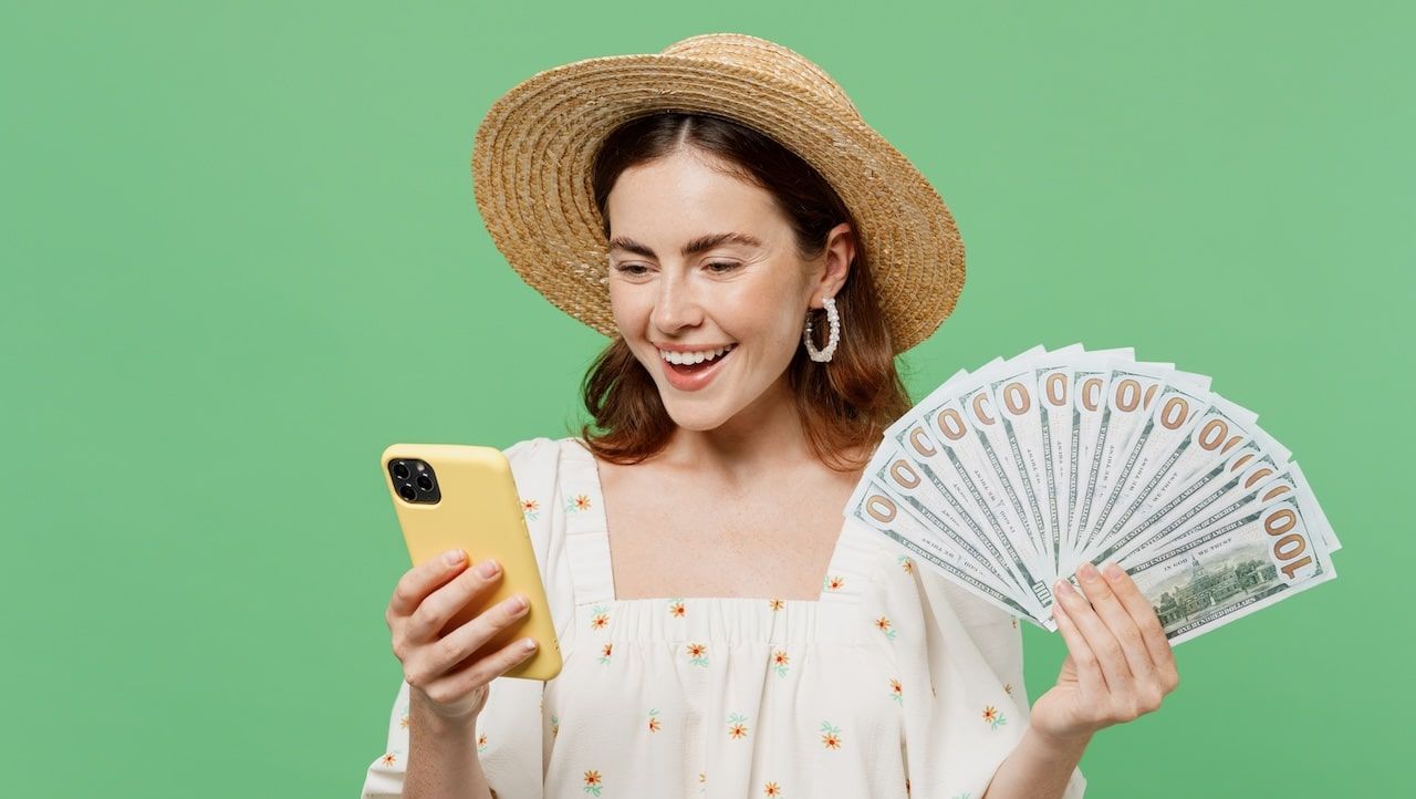 Young happy smiling woman she 20s in white dress hat hold in hand use mobile cell phone fan of cash money in dollar banknotes isolated on plain pastel light green background.
