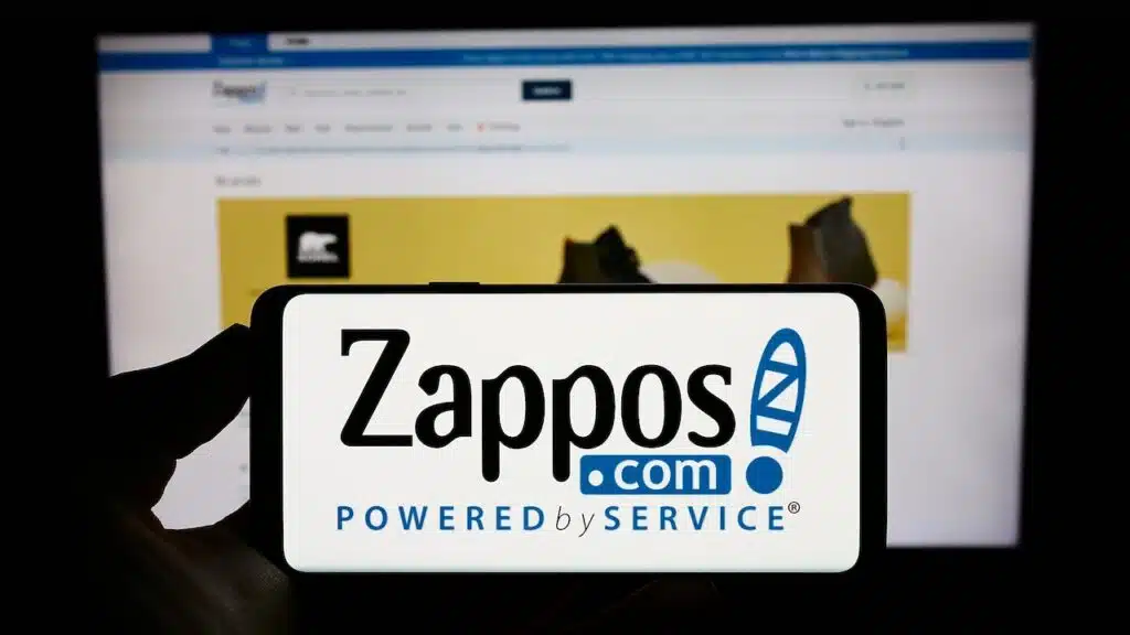 Person holding cellphone with logo of US online shop company Zappos.com LLC on screen in front of business webpage.