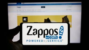 Person holding cellphone with logo of US online shop company Zappos.com LLC on screen in front of business webpage.