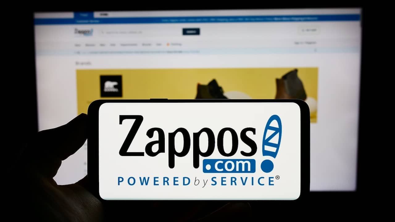 Person holding cellphone with logo of US online shop company Zappos.com LLC on screen in front of business webpage.