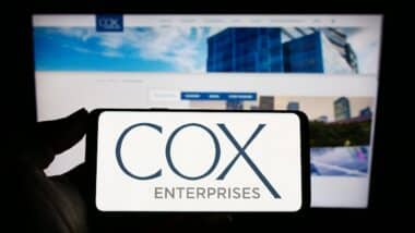 Person holding mobile phone with logo of American company Cox Enterprises Inc. on screen in front of business web page.