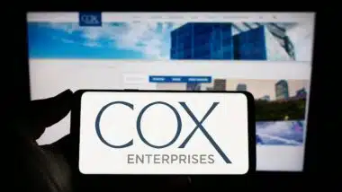 Person holding mobile phone with logo of American company Cox Enterprises Inc. on screen in front of business web page.