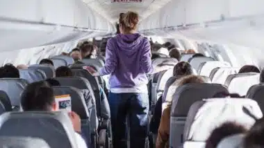 Interior of airplane with passengers on seats and female traveler walking the aisle.