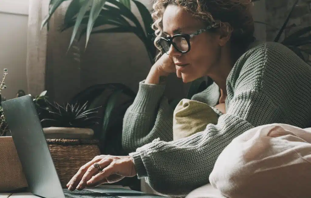 Blonde woman with curly hair using laptop sitting on sofa at home.