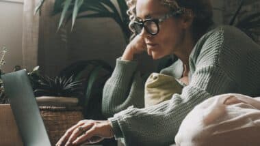 Blonde woman with curly hair using laptop sitting on sofa at home.