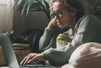 Blonde woman with curly hair using laptop sitting on sofa at home.