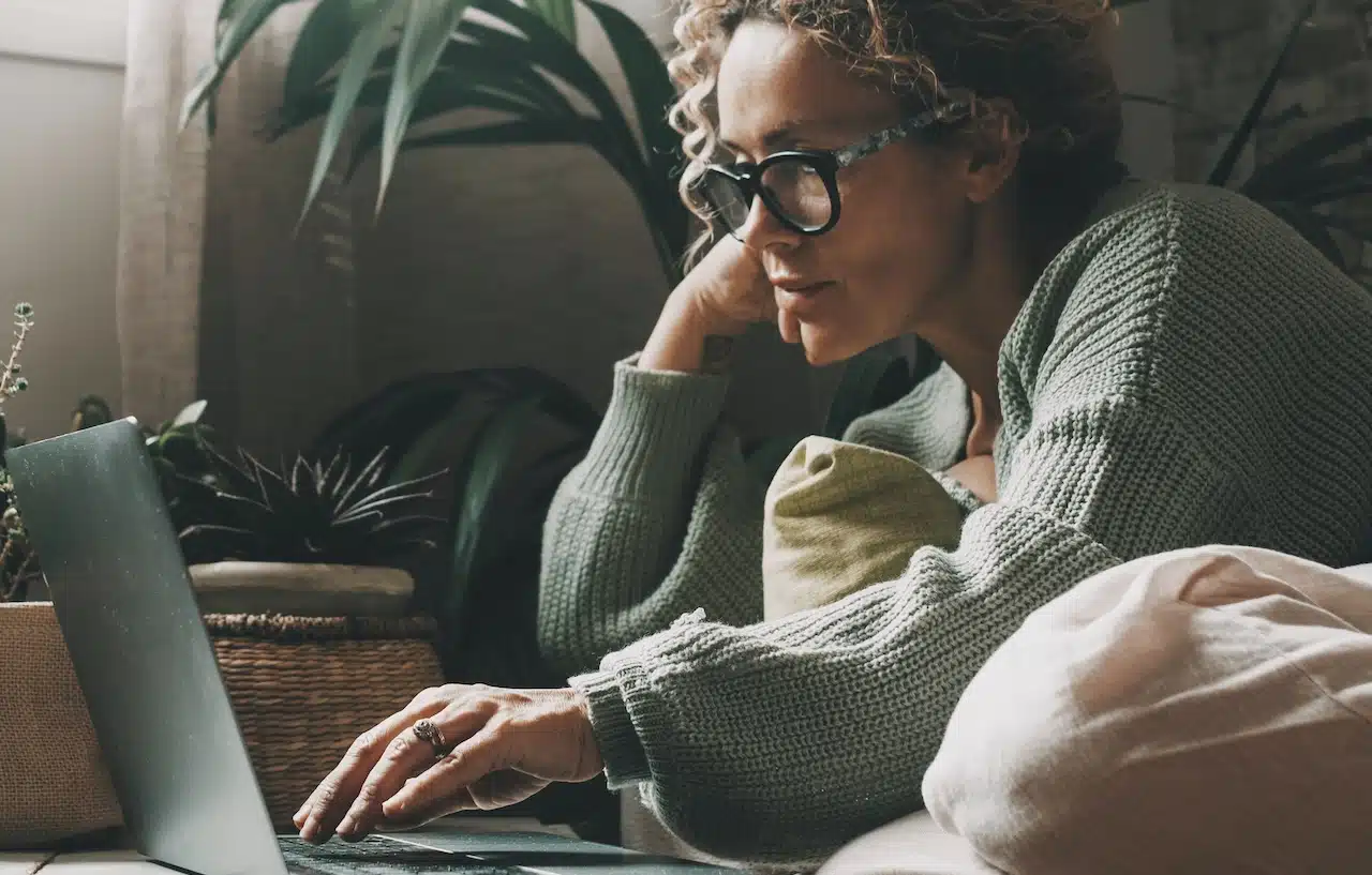 Blonde woman with curly hair using laptop sitting on sofa at home.