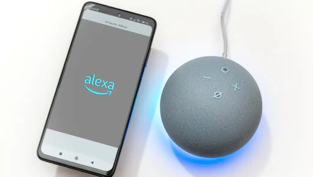 Hand holding a smartphone with Alexa logo next to an Echo Dot smart speaker on a white background with the blue light ring activated and showing the time