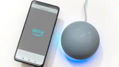 Hand holding a smartphone with Alexa logo next to an Echo Dot smart speaker on a white background with the blue light ring activated and showing the time