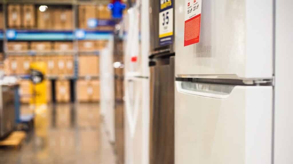 Row of refrigerators in Dallas appliance store with visible price tags and polished floor. Blurry background of shelves, boxes, forklift pallet mover in warehouse setting, home improvement store.