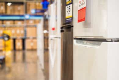 Row of refrigerators in Dallas appliance store with visible price tags and polished floor. Blurry background of shelves, boxes, forklift pallet mover in warehouse setting, home improvement store.