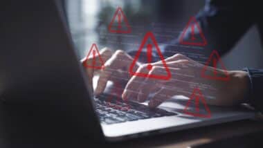 Cybersecurity Alert: Hands Typing on Laptop with Overlay of Code and Multiple Red Warning Signs Indicating System Error or Data Breach Threat