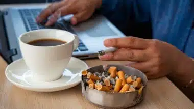 Focus on many cigarette butts in a fulled and dirty ashtray. A stressed woman smoking cigarette drinking black coffee working under pressure from home on laptop as the background.
