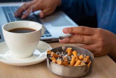 Focus on many cigarette butts in a fulled and dirty ashtray. A stressed woman smoking cigarette drinking black coffee working under pressure from home on laptop as the background.