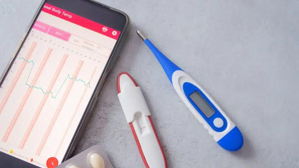 Fertility tracking tools, including a BBT thermometer, an app displaying a temperature chart, and supplements for pregnancy planning.