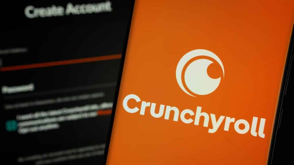 Crunchyroll logo is displayed on a smartphone screen, with the crunchyroll login page visible on a computer screen in the background.
