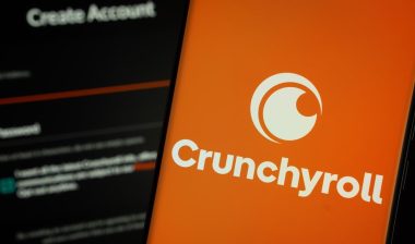 Crunchyroll logo is displayed on a smartphone screen, with the crunchyroll login page visible on a computer screen in the background.