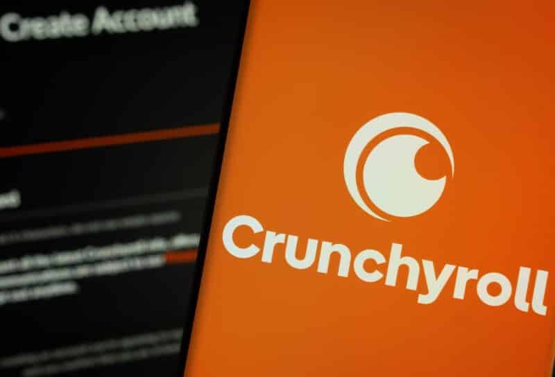 Crunchyroll logo is displayed on a smartphone screen, with the crunchyroll login page visible on a computer screen in the background.