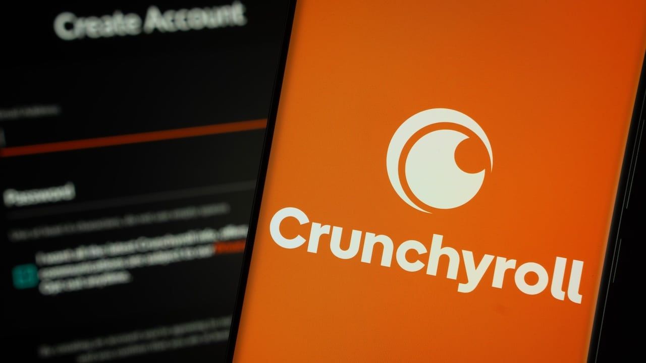 Crunchyroll logo is displayed on a smartphone screen, with the crunchyroll login page visible on a computer screen in the background.