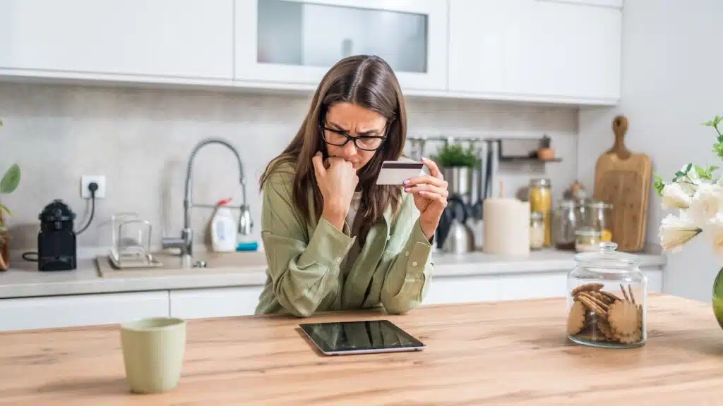 holding a credit card and staring at a tablet concerned about fraud, hidden fees, and tight budgeting while shopping from home