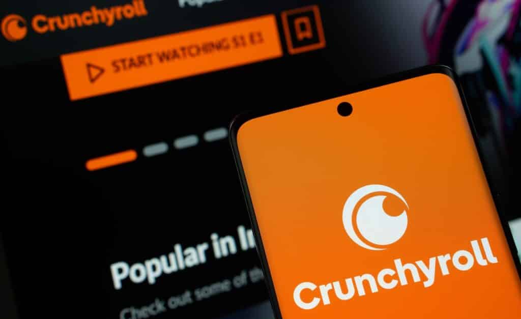 Crunchyroll logo is displayed on a smartphone screen, with the crunchyroll website visible on a computer screen in the background.