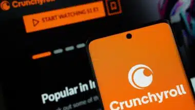 Crunchyroll logo is displayed on a smartphone screen, with the crunchyroll website visible on a computer screen in the background.