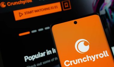 Crunchyroll logo is displayed on a smartphone screen, with the crunchyroll website visible on a computer screen in the background.