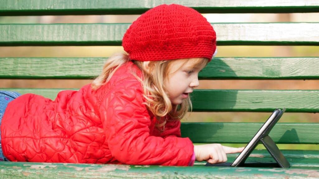 Happy smiling little girl working with tablet pc and lying on bench in the park.