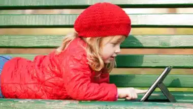 Happy smiling little girl working with tablet pc and lying on bench in the park.